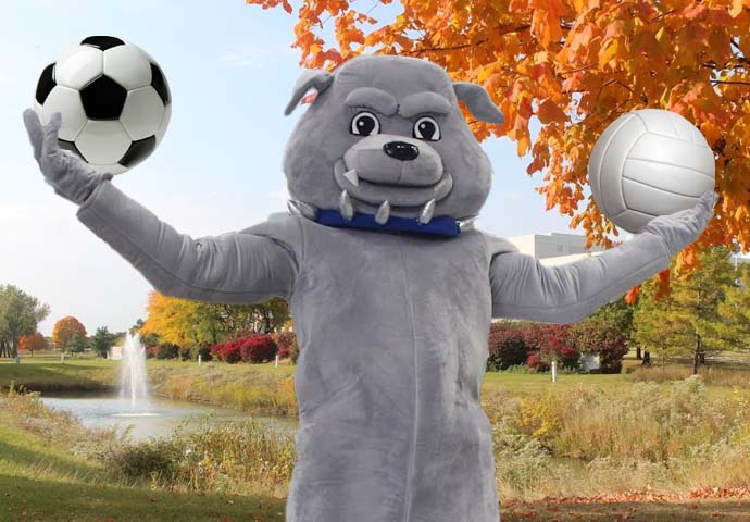 Bulldog mascot holding a soccer and volleyball in front of fall trees.